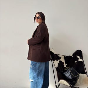May include: A woman wearing a brown blazer, blue jeans with distressed details, and sunglasses. She is standing in front of a white wall with a black and white cowhide chair in the background.