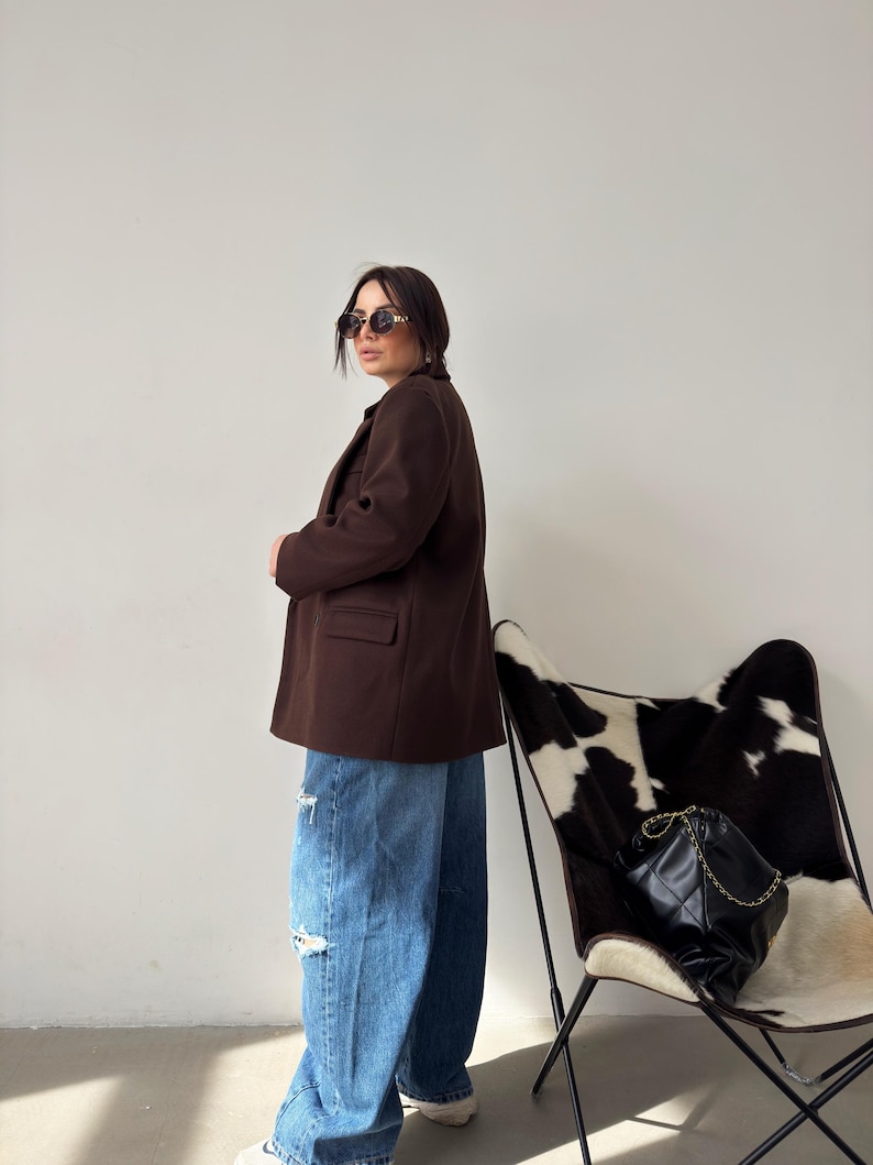 May include: A woman wearing a brown blazer, blue jeans with distressed details, and sunglasses. She is standing in front of a white wall with a black and white cowhide chair in the background.