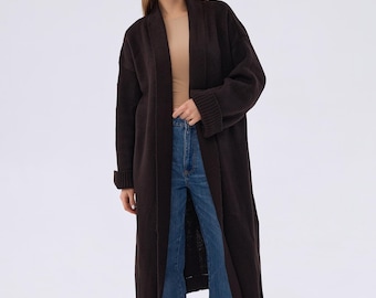 May include: A long brown cardigan with a wide collar and long sleeves. The cardigan is open in the front and the model is wearing it over a light brown top and blue jeans.