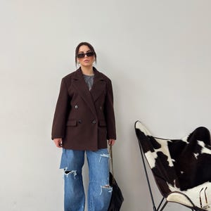 May include: A woman wearing a brown double-breasted blazer, blue jeans with distressed details, and white trainers. She is carrying a black handbag and wearing sunglasses.
