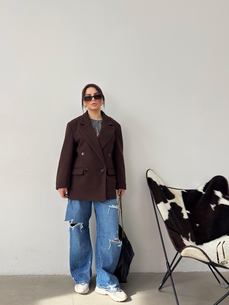 May include: A woman wearing a brown double-breasted blazer, blue jeans with distressed details, and white trainers. She is carrying a black handbag and wearing sunglasses.