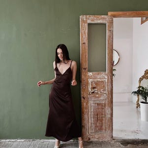 K&ouml;nnte beinhalten: Eine Frau in einem braunen Satin-Maxikleid und wei&szlig;en Sandaletten mit Absatz steht vor einer gr&uuml;nen Wand und einer Holzt&uuml;r.