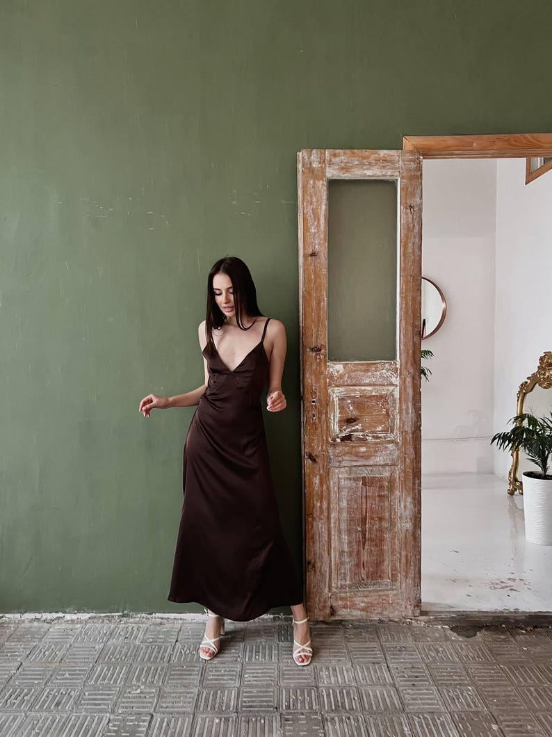 K&ouml;nnte beinhalten: Eine Frau in einem braunen Satin-Maxikleid und wei&szlig;en Sandaletten mit Absatz steht vor einer gr&uuml;nen Wand und einer Holzt&uuml;r.