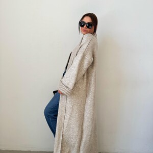 May include: A long, beige cardigan with a shawl collar and long sleeves. The cardigan is open in the front and has a relaxed fit.