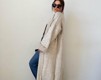 May include: A long, beige cardigan with a shawl collar and long sleeves. The cardigan is open in the front and has a relaxed fit.