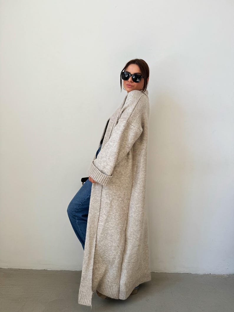 May include: A long, beige cardigan with a shawl collar and long sleeves. The cardigan is open in the front and has a relaxed fit.