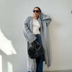 May include: A grey long-sleeved cardigan with a white ribbed tank top underneath. The person is wearing blue jeans and black sunglasses. They are holding a black quilted handbag with a gold chain strap.