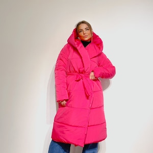 May include: A vibrant fuchsia puffer coat with a large hood and a matching belt. The long coat is worn over blue jeans. The coat has a quilted design and a wrap-style closure. The model is wearing a black turtleneck.