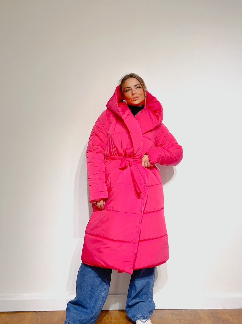 May include: A vibrant fuchsia puffer coat with a large hood and a matching belt. The long coat is worn over blue jeans. The coat has a quilted design and a wrap-style closure. The model is wearing a black turtleneck.