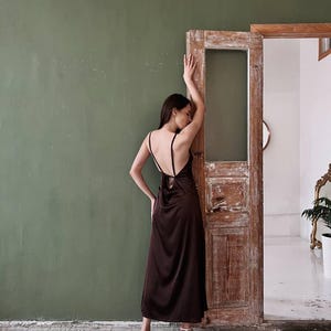 K&ouml;nnte beinhalten: Eine Frau in einem braunen Satin-Maxikleid mit tiefem R&uuml;cken und einem Bindedetail. Sie steht vor einer Holzt&uuml;r mit einer gr&uuml;nen Wand dahinter.