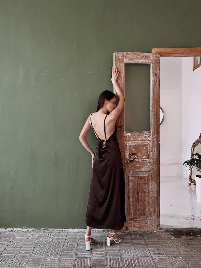 K&ouml;nnte beinhalten: Eine Frau in einem braunen Satin-Maxikleid mit tiefem R&uuml;cken und einem Bindedetail. Sie steht vor einer Holzt&uuml;r mit einer gr&uuml;nen Wand dahinter.