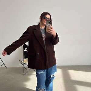 May include: A woman wearing a brown double-breasted blazer over a grey t-shirt with a graphic print. She is wearing blue jeans with rips and white trainers.