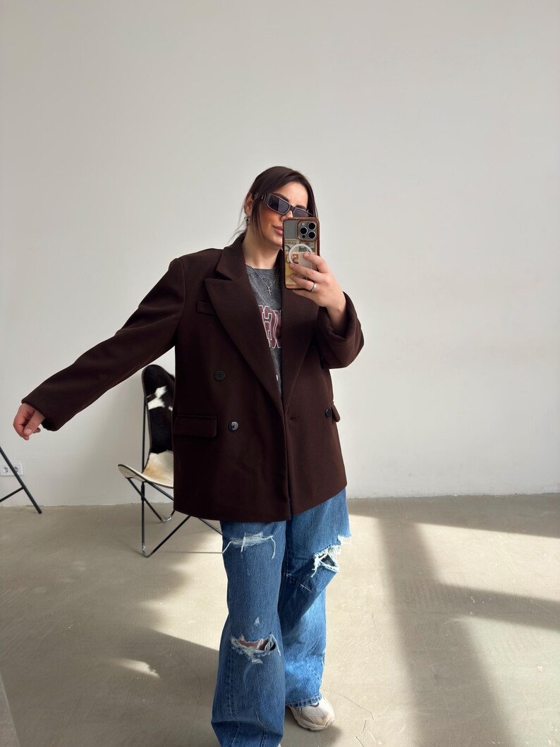 May include: A woman wearing a brown double-breasted blazer over a grey t-shirt with a graphic print. She is wearing blue jeans with rips and white trainers.