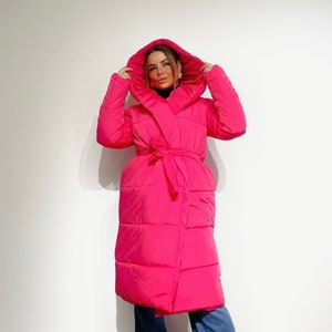 May include: A bright pink, hooded puffer coat with a wrap-around design and a matching tie belt. The long coat features a quilted pattern and is paired with blue jeans. The model is wearing a black turtleneck.