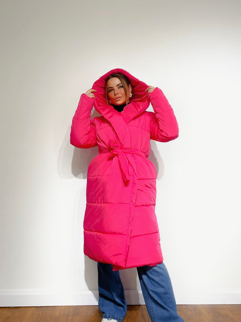 May include: A bright pink, hooded puffer coat with a wrap-around design and a matching tie belt. The long coat features a quilted pattern and is paired with blue jeans. The model is wearing a black turtleneck.
