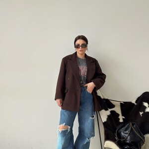 May include: A woman wearing a brown blazer, a grey t-shirt with the word "Angel" printed on it, and blue jeans with distressed details. She is standing in front of a white and black cowhide chair.