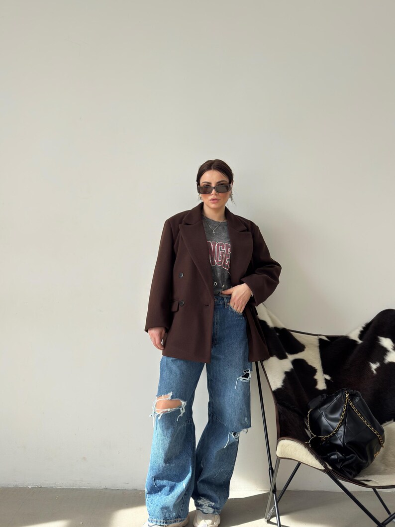 May include: A woman wearing a brown blazer, a grey t-shirt with the word "Angel" printed on it, and blue jeans with distressed details. She is standing in front of a white and black cowhide chair.