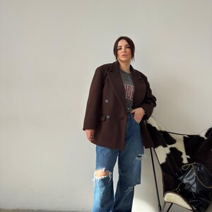 May include: A woman wearing a brown double-breasted blazer over a grey t-shirt with a red and white graphic print. She is wearing blue jeans with distressed details and white trainers.
