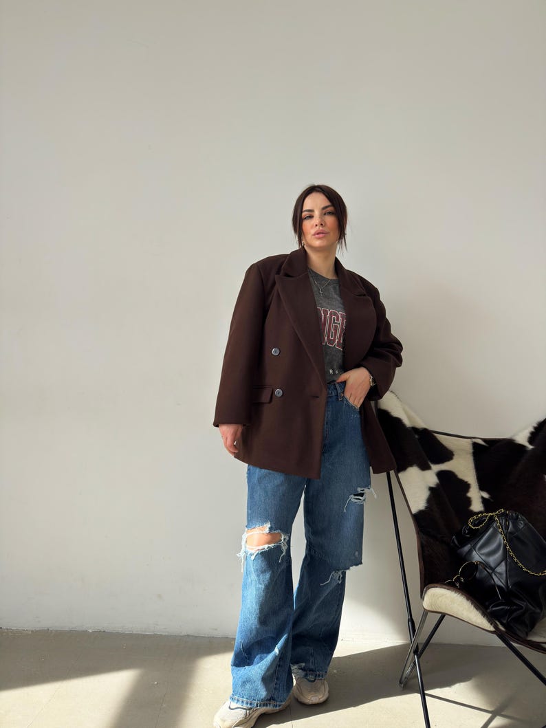 May include: A woman wearing a brown double-breasted blazer over a grey t-shirt with a red and white graphic print. She is wearing blue jeans with distressed details and white trainers.