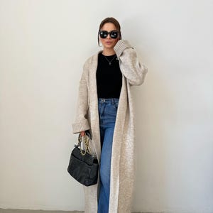May include: A woman wearing a long, beige cardigan with a black top and blue jeans. She is carrying a black handbag with a gold chain strap.