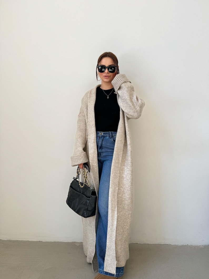 May include: A woman wearing a long, beige cardigan with a black top and blue jeans. She is carrying a black handbag with a gold chain strap.
