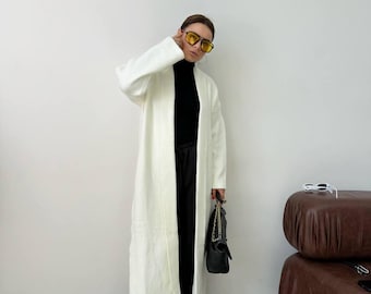 Op de afbeelding: Een witte, langarmige cardigan met een zwarte turtleneck eronder. De cardigan is open aan de voorkant en heeft een lange, vloeiende silhouet. De persoon draagt een zwarte broek en bruine schoenen.