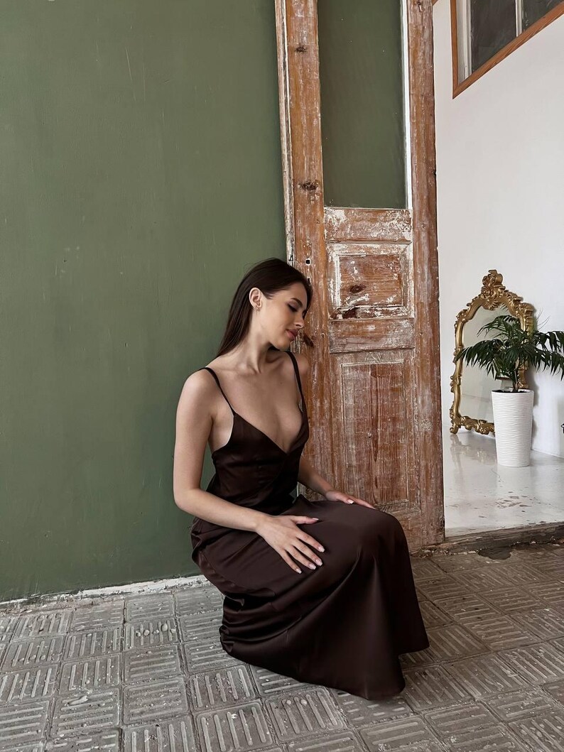 K&ouml;nnte beinhalten: Eine Frau in einem braunen Satin-Maxikleid, die auf einem gefliesten Boden sitzt, mit einer gr&uuml;nen Wand und einer Holzt&uuml;r im Hintergrund.