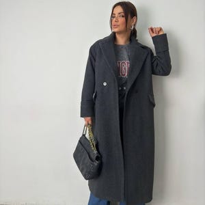 May include: A charcoal gray double-breasted overcoat with large lapels. The coat is worn over a gray t-shirt with the word "ING" in maroon lettering. A black quilted handbag with a gold chain strap is held in the left hand. The person is wearing blue jeans and tan shoes.