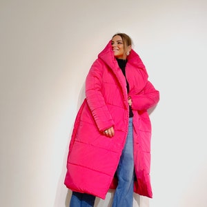 May include: A bright pink, long puffer coat with a large hood. The coat is worn over a black top and blue jeans. The coat has a belt and is knee-length.