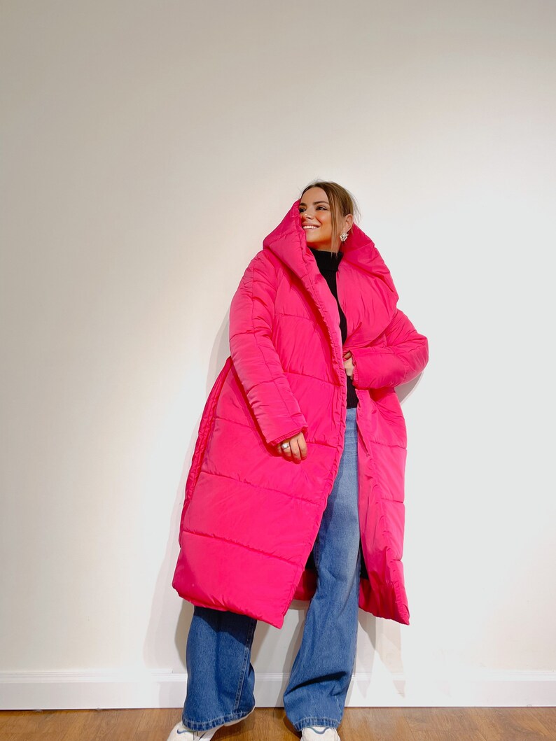 May include: A bright pink, long puffer coat with a large hood. The coat is worn over a black top and blue jeans. The coat has a belt and is knee-length.