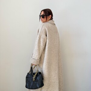 May include: A long, beige cardigan with a collar and long sleeves. The cardigan is made of a soft, knitted fabric. The person is wearing black sunglasses and a black handbag with a gold chain.