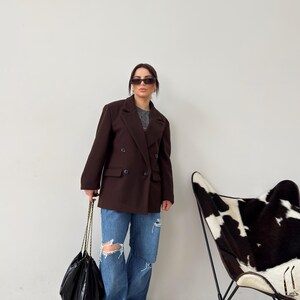 May include: A woman wearing a brown double-breasted blazer, blue jeans with distressed details, and black sunglasses. She is carrying a black chain bag. A black and white cowhide patterned chair is in the background.