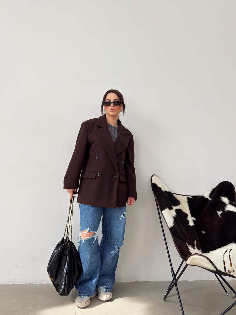 May include: A woman wearing a brown double-breasted blazer, blue jeans with distressed details, and black sunglasses. She is carrying a black chain bag. A black and white cowhide patterned chair is in the background.