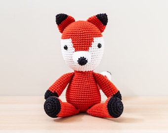 May include: A crocheted fox plush toy with orange fur, black ears, and black eyes. The toy is sitting on a wooden surface.