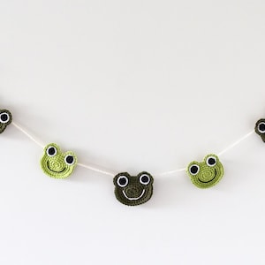 May include: A green and brown crocheted frog garland with black eyes. The garland is strung on white string and is perfect for a nursery or child's room.