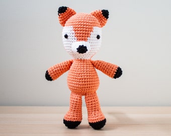 May include: A crocheted orange fox plush toy with black ears, black eyes, and a black nose. The toy is standing on a wooden surface.