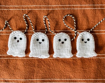 May include: A string of five white crocheted ghosts with black eyes and mouths hanging on a brown and white striped fabric background.