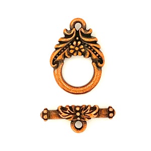 May include: Copper-toned metal clasp with a floral design. The clasp has a ring and a bar with a decorative floral design.