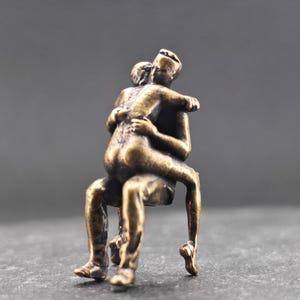 May include: A small bronze-coloured metal figurine depicting two figures in an embrace. One figure is seated while the other sits on their lap, with arms wrapped around the seated figure's neck. The sculpture has a textured, weathered finish.