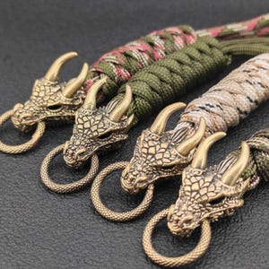 May include: Close-up of four dragon head keychains. The dragon heads are bronze-colored with intricate detailing and attached to a braided cord. The cords are in shades of green, brown, and camouflage patterns.