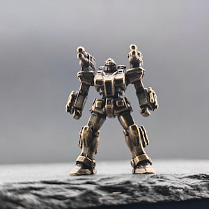 May include: A small, metallic bronze-colored figurine of a humanoid robot standing in a bipedal stance. The robot features two shoulder-mounted cannons, articulated limbs, and a detailed mechanical torso with a weathered, antique finish.