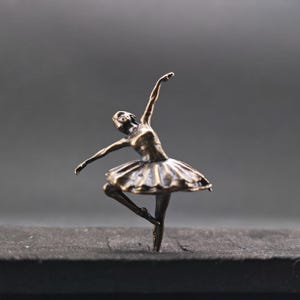 Könnte beinhalten: Eine kleine bronzefarbene Figur einer tanzenden Person in Ballettpose. Die Figur ist in einer Bewegung mit einem nach oben und einem zur Seite gestreckten Arm dargestellt, trägt einen plissierten Tutu-Rock und steht auf einer Fußspitze.