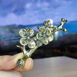 May include: A bronze floral branch featuring three detailed flowers and a spiral design. The sculpture is held up, highlighting the intricate details of the blossoms and stem. The background is blurred, focusing attention on the metalwork.