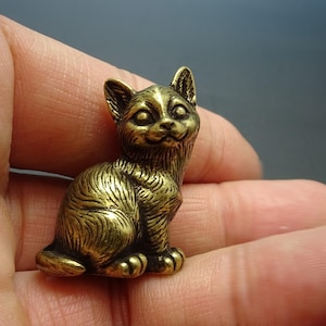 May include: A small, gold-coloured brass figurine of a cat sitting with its head tilted up. The cat has a detailed, textured fur pattern and large, round eyes.
