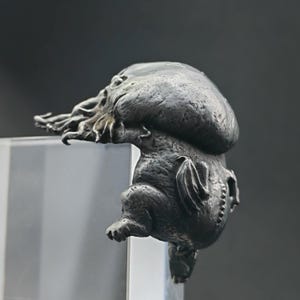 May include: A detailed, silver-coloured sculpture of a creature with a bulbous head, tentacles, and bat-like wings. The sculpture has a textured surface and is perched on a clear, rectangular block. The creature's design is reminiscent of a mythical being.