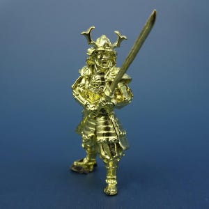 May include: A gold-coloured figurine of a samurai warrior holding a sword. The warrior is wearing traditional armour and a helmet with horns.