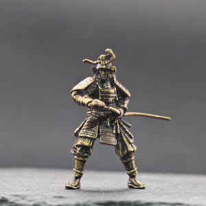 May include: A small, antique-style bronze-colored metal figurine of a samurai warrior in traditional armor. The figure stands in a defensive stance, holding a katana sword with both hands, featuring detailed plate armor, a helmet, and textured finish.