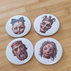Puede incluir: Cuatro chapas redondas con ilustraciones detalladas de cabezas de muñecos. Una muestra un bebé con un lazo azul, otra un bufón con corona, otra un rey barbudo con textura de madera y otra una muñeca con pelo revuelto y cara cosida.