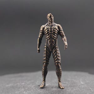 Könnte beinhalten: Eine bronzefarbene Figur eines menschlichen Skeletts. Die Figur steht aufrecht mit den Armen an den Seiten und zeigt detaillierte Skelett- und Muskelstrukturen. Die Figur hat ein verwittertes Aussehen, was auf eine antike oder Vintage-Ästhetik hindeutet.