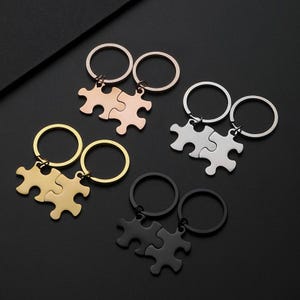 May include: A collection of matching puzzle piece keychains in gold, rose gold, silver, and black. Each keychain set features two interlocking puzzle pieces attached to a metal ring. The keychains are displayed on a dark surface.